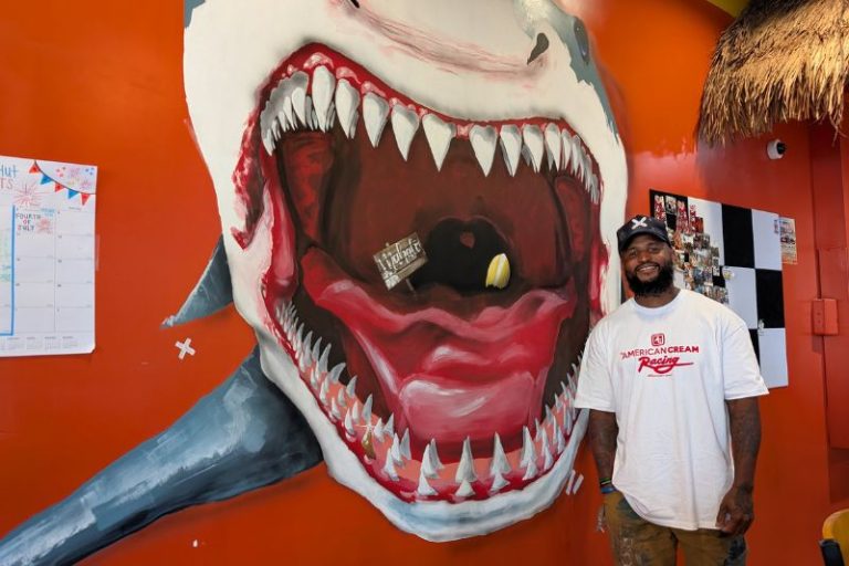 `Artist "Mammxth" stands in front of one of 2 murals he painted at Beach Hut Deli. // SB WIlliams
